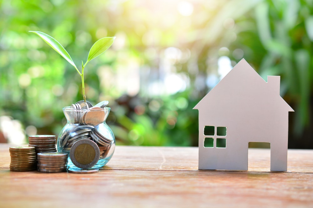 A jar of coins with a plant sprouting out of it beside a wood house model.