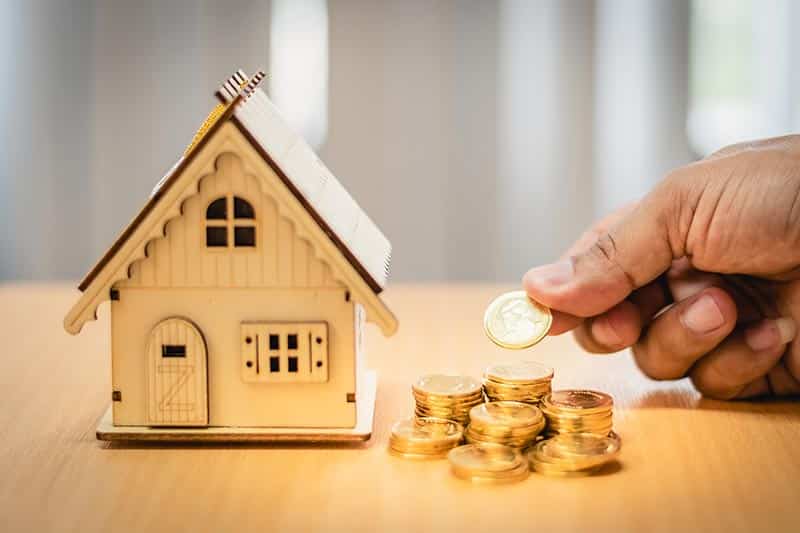 A hand placing coins next to a tiny, wooden house.