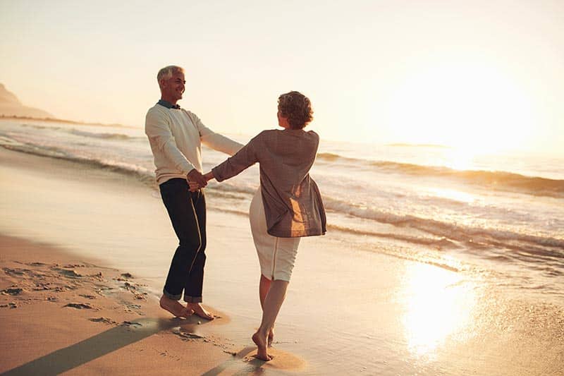 A 55+ couple with arthritis holding hands and enjoying a day at the beach.