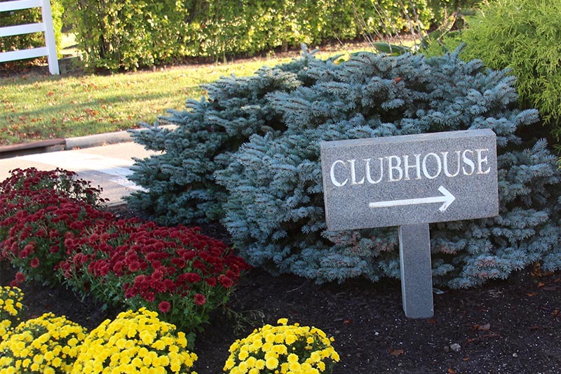 Sign near shrubs and plants that says clubhouse with a right facing arrow underneath the words.