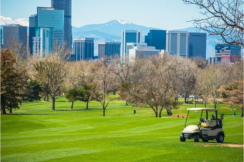 Golf cart on tree lined course with Denver skyline and Rocky Mountains in the background.