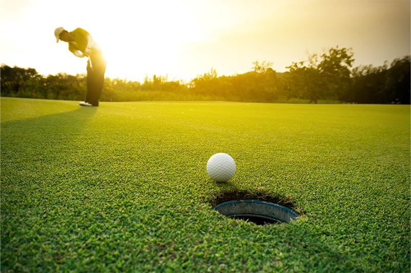 Man putting golf ball at edge of hole at sunrise.