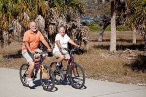 Older couple riding bikes along a path beside palm trees and dry grass.