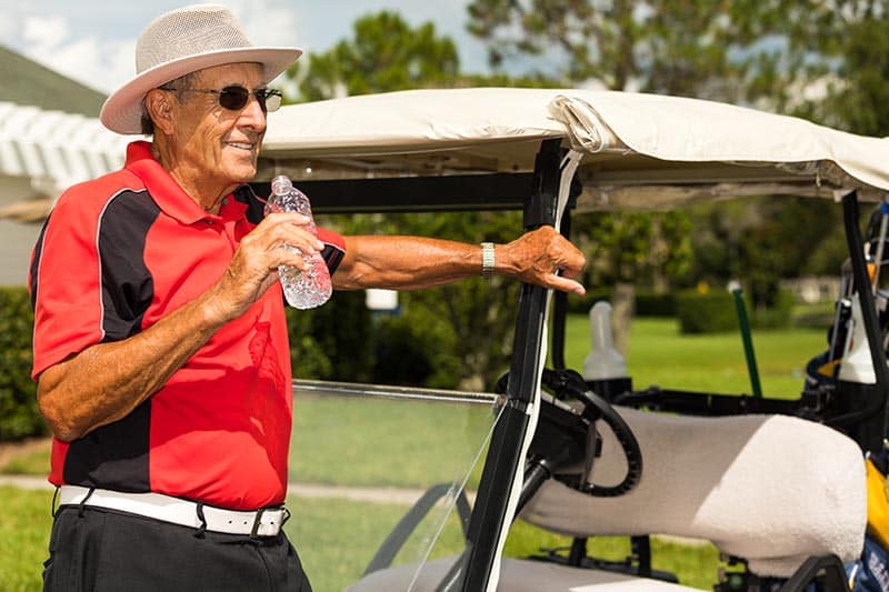 Senior man leaning against golf cart while drinking a bottle of water.