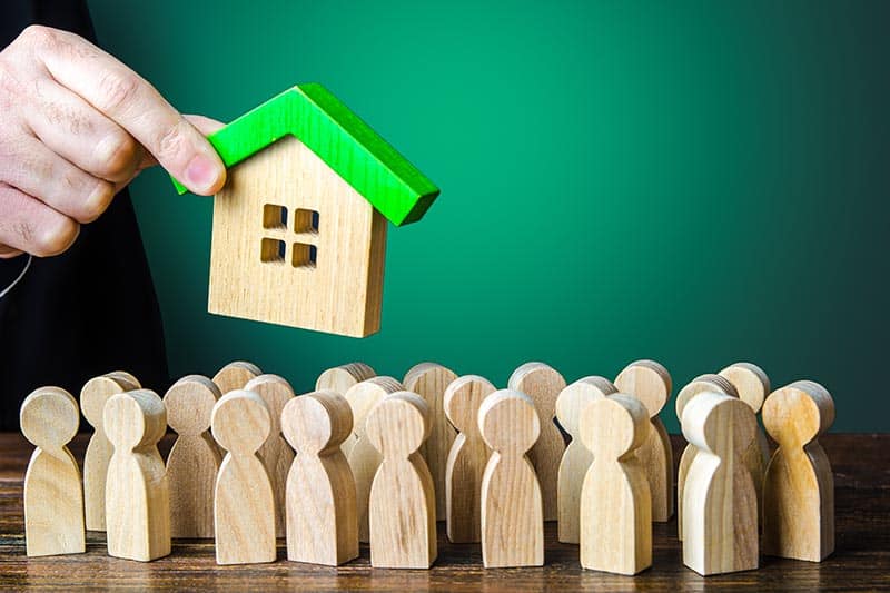 Man's hand holding a wooden house over a group of wooden figures.