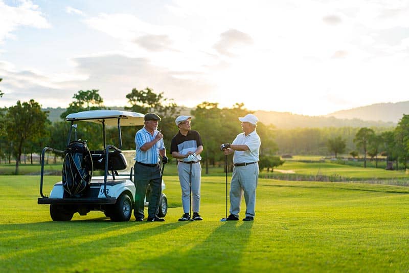 Three older men on golf course standing near a golf cart chatting.