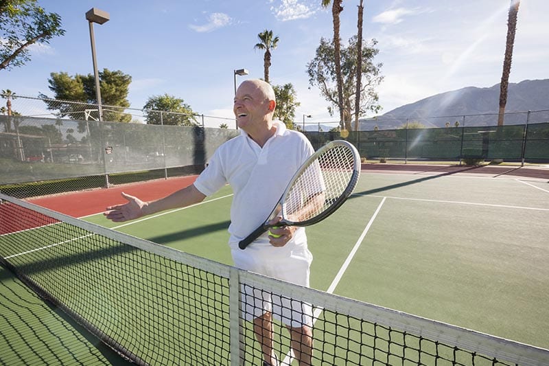 Older man holding tennis racket and reaching his hand out for a handshake over the tennis net.