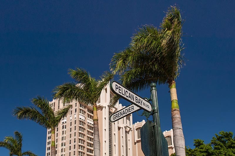 Pelican Bay decorative street signs with tall building, palm trees and blue sky in the background.