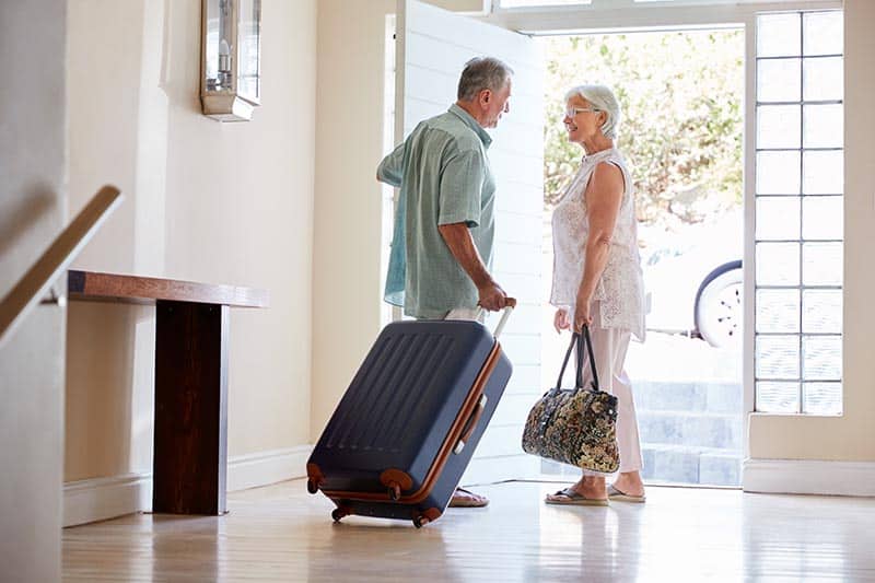 Senior couple standing by front door with suitcase about to leave for vacation.