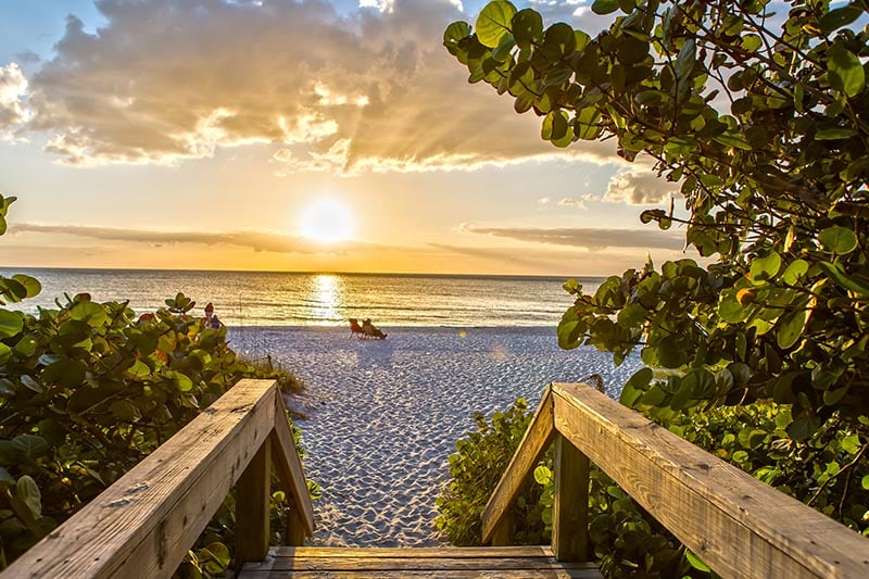 Stair leading down to beach at sunset, couple sitting in the sand in the distance.