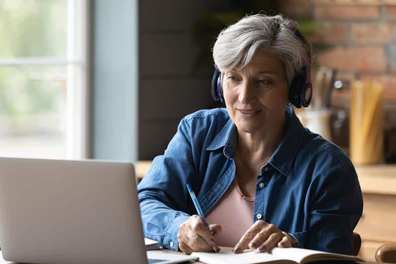 A 55+ woman sitting at a table wear headphones and taking notes on a laptop.