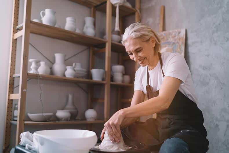 Smiling senior woman molding clay pot on pottery wheel in art studio.