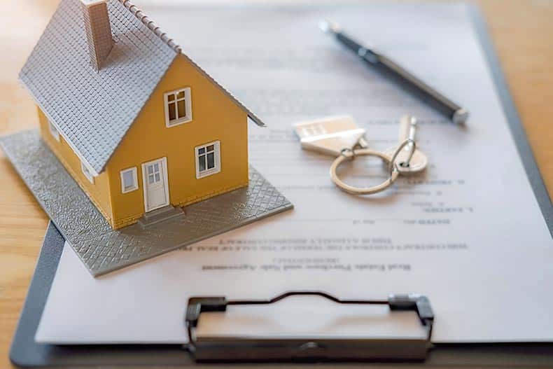 A tiny model of a house on top of a real estate contract beside keys and a pen.