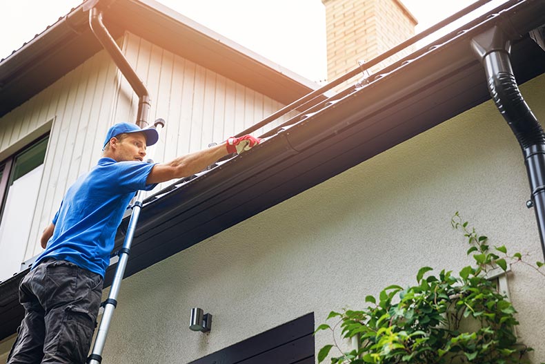 A man on a ladder cleaning a house gutter from leaves and dirt.