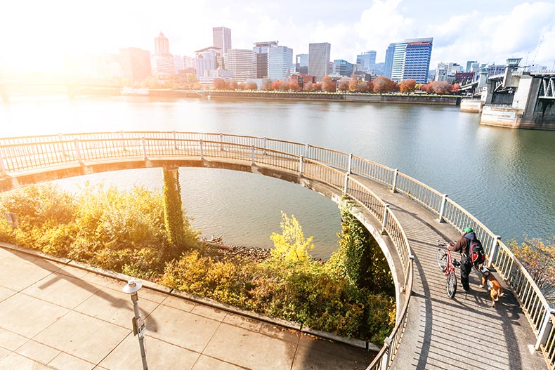 A person with a bicycle walking on an elevated bridge near the water with a view of the Portland skyline in Oregon.