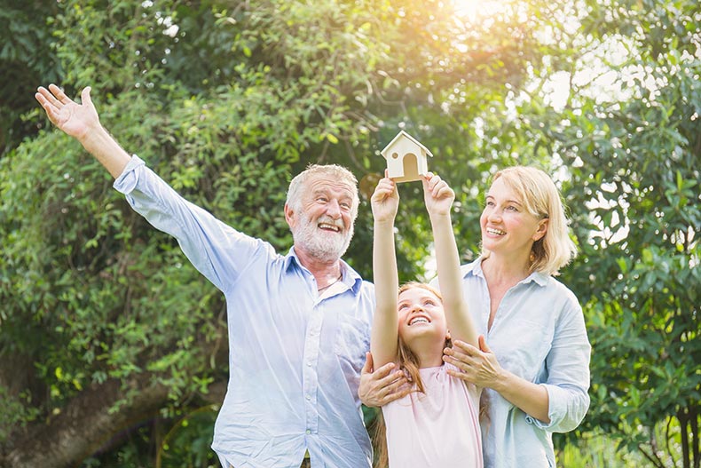 A happy family with a 55+ man, a woman, and a little girl holding a model house.