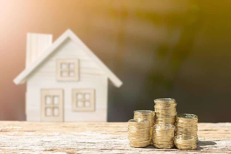 A stack of coins in front of a wooden model house on a table.