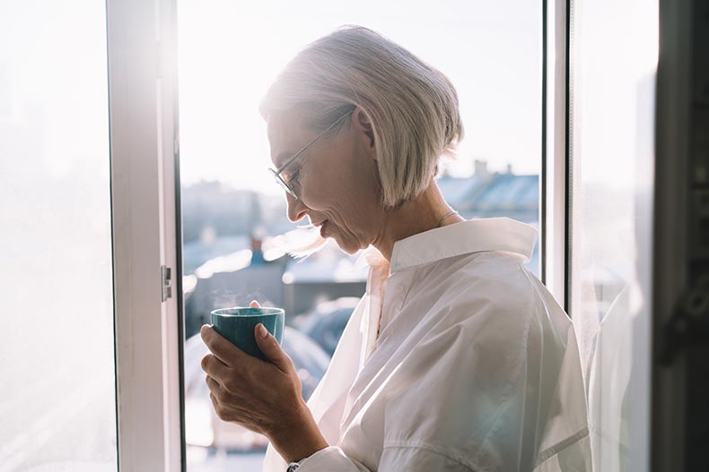 A mature woman drinking coffee from mug near a window with a view of the city.