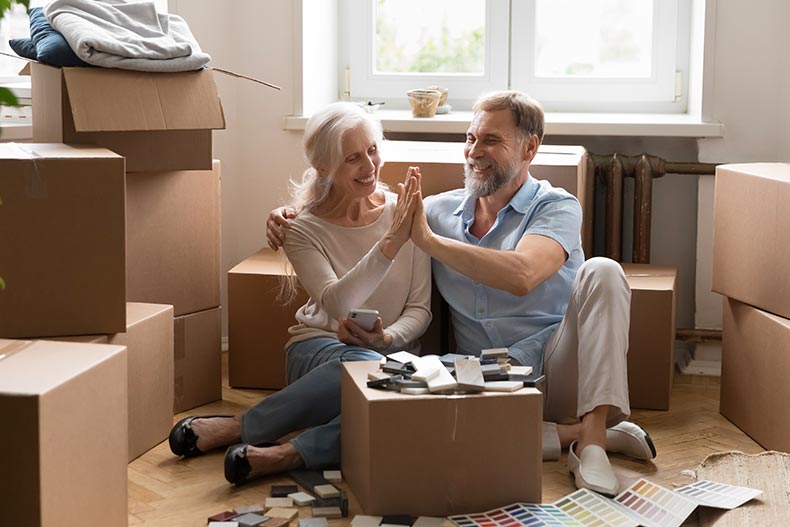 Older spouses smiling and sitting on the floor near a heap of cardboard boxes.