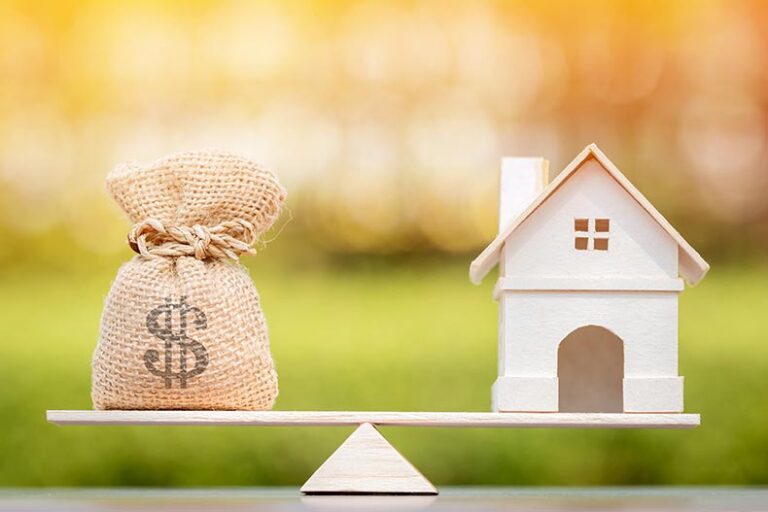 A bag of money balancing on a seesaw with a wood house model.