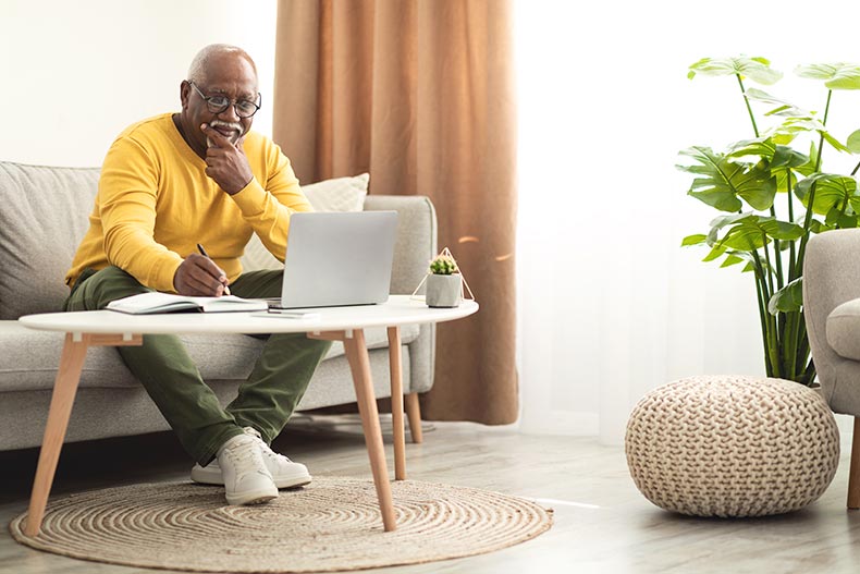 A man taking notes while using a laptop and sitting on a sofa at home.