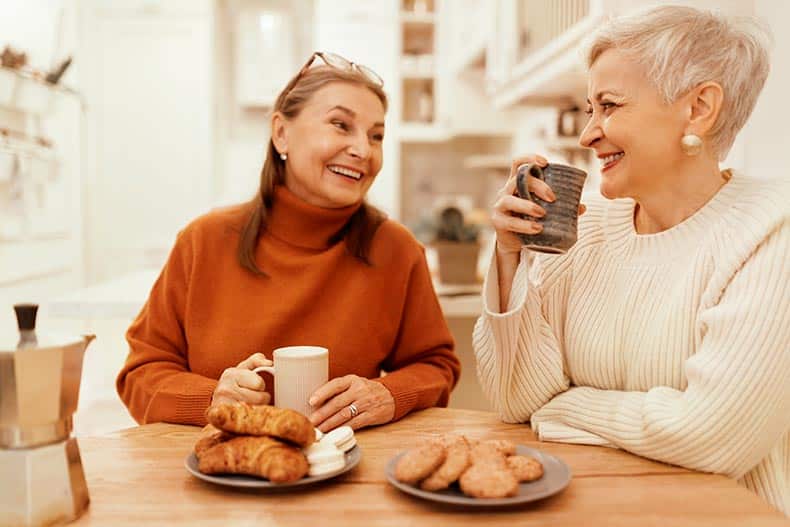 Two stylish senior women enjoying warm beverages and sweets together.