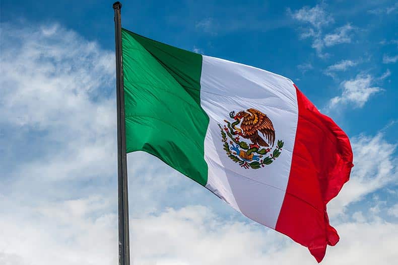 The Mexico flag flying with a blue sky with white clouds behind it.