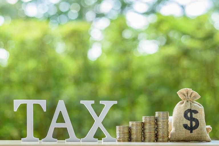 The word tax beside rows of rising coins and a U.S. dollar bag on a table.