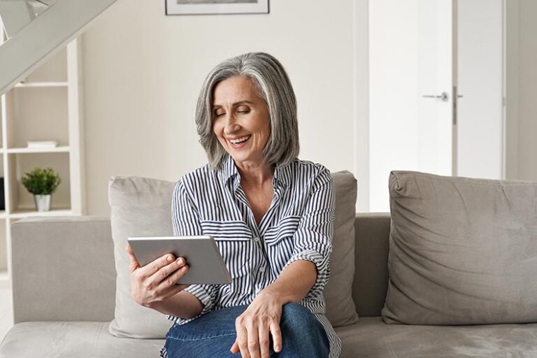 A mature woman smiling while sitting on the couch and using an app to help her sell her house.