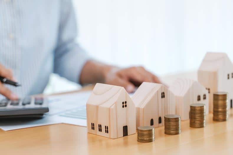 A row of wooden home models and stacks of coins beside a man calculating his property taxes.