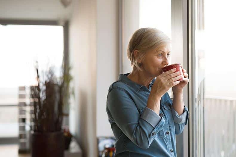 A senior woman drinking a cup of coffee while looking out her apartment window.