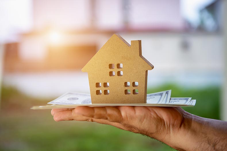 A hand holding a wooden house model on top of a stack of cash.