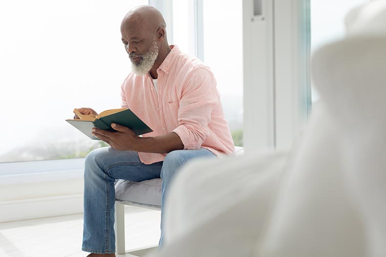 A 55+ man reading a book while sitting on the bed in his apartment.