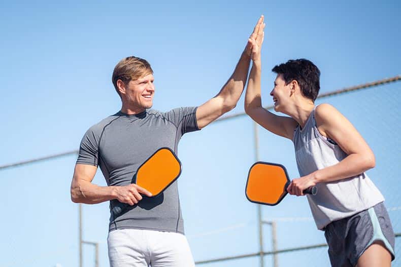 A 55+ couple laughing and high-fiving during a pickleball game.