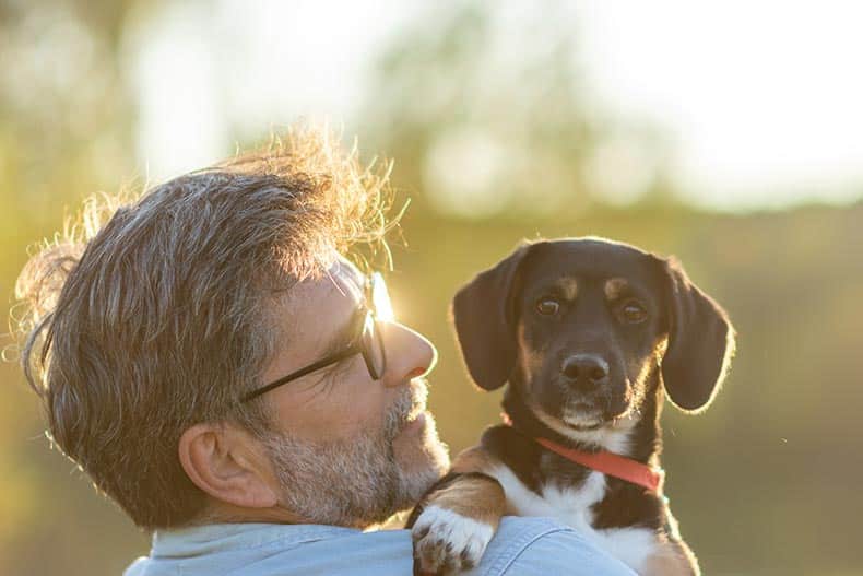 A 55+ man with his dog hugging and playing outdoors in the park.