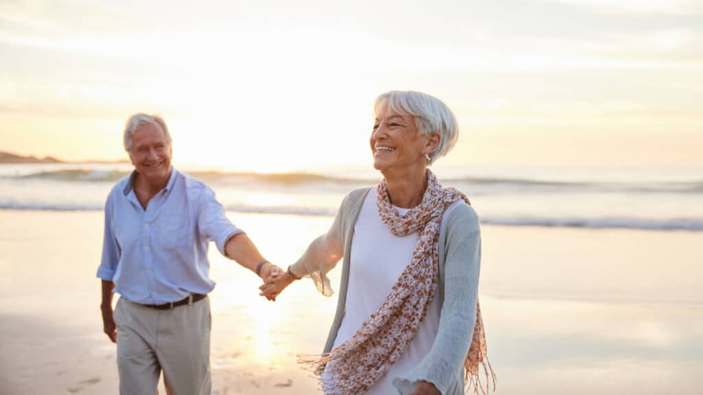 Senior man and woman hold hands on the beach, choosing to move out-of-state for retirement.