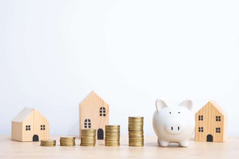 Wooden house models behind stacks of coins and a piggy bank.