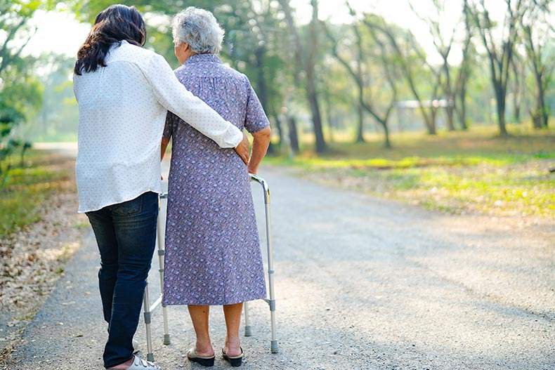 A woman helping her mother walk down a nature path with a walker.