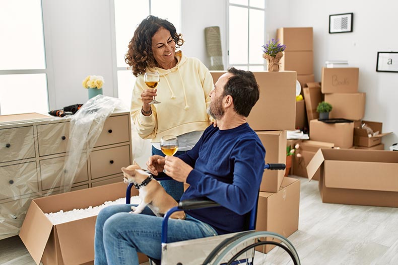 A 55+ couple toasting with champagne as the man sits in a wheelchair in their new home.