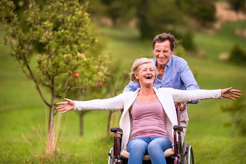 A 55+ man pushing a 55+ woman in a wheelchair while both smile and laugh.