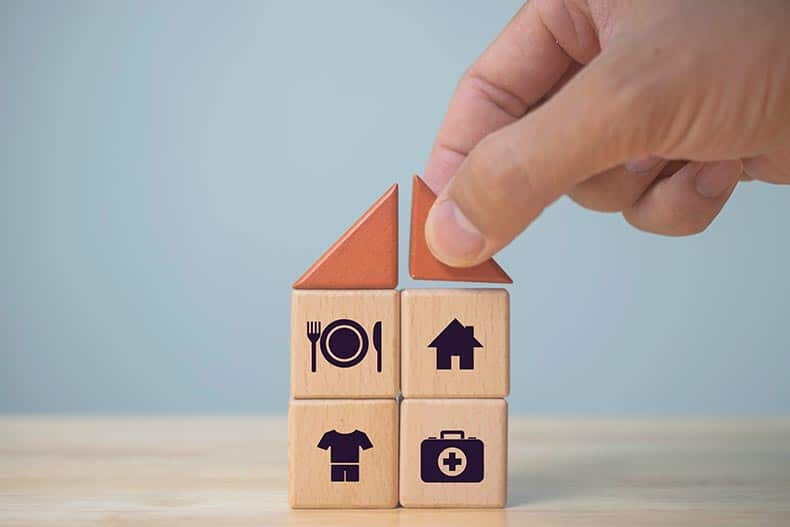 A hand building wooden cubes with icons representing food, clothing, housing, and medicine.