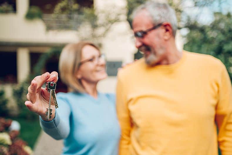 A 55+ couple holding keys and standing outside their new home on a moving day.