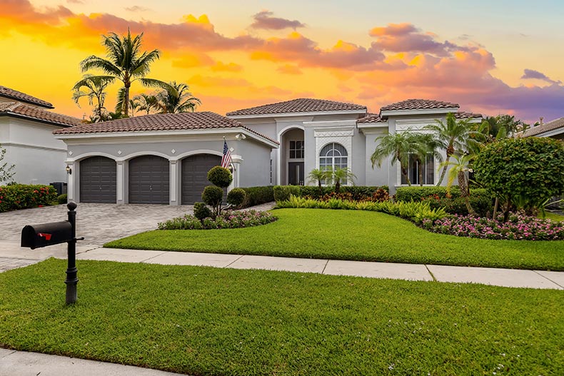 An elegant house with gray walls, a red tiled roof, a front garden with abundant tropical plants, a driveway, and a garage.