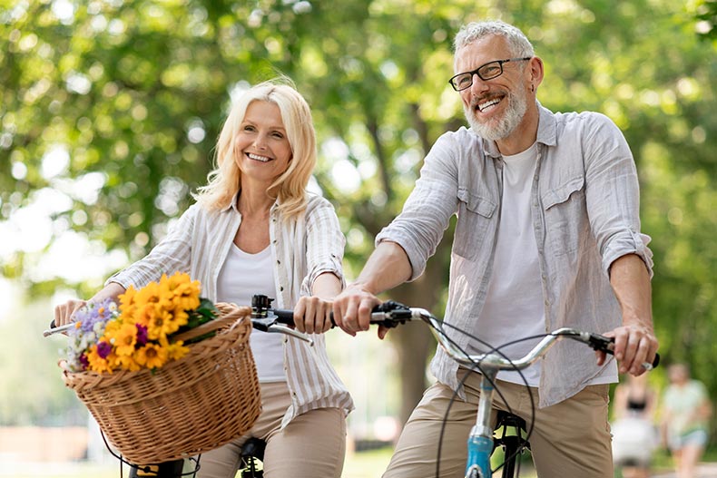 A 55+ couple smiling while riding bikes in a park on a sunny day.