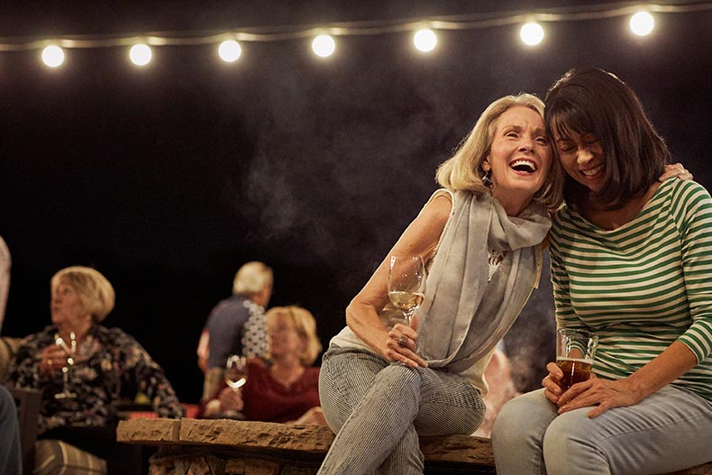 A group of active adults laughing and drinking around the outdoor fire pit in their 55+ community.