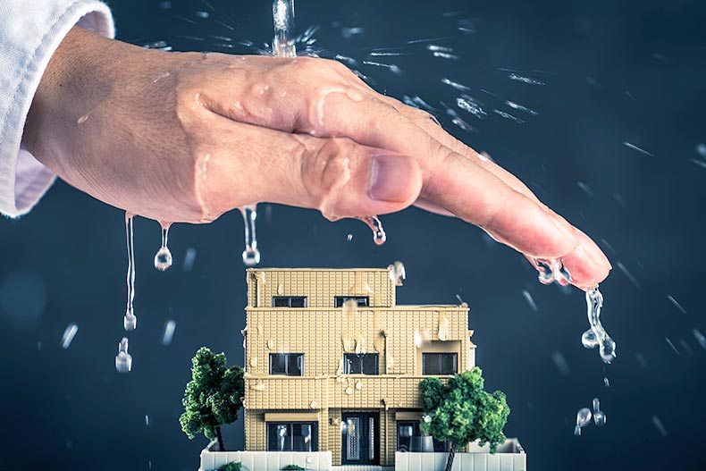 Closeup of a hand over a model house preventing water from destroying it.