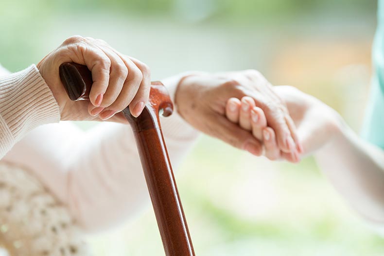 Closeup of an elderly lady holding a walking cane in one hand and holding a nurse's hand in the other.