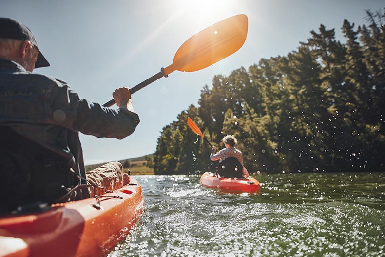 A senior man canoeing in the lake with a woman in background on a summer day.