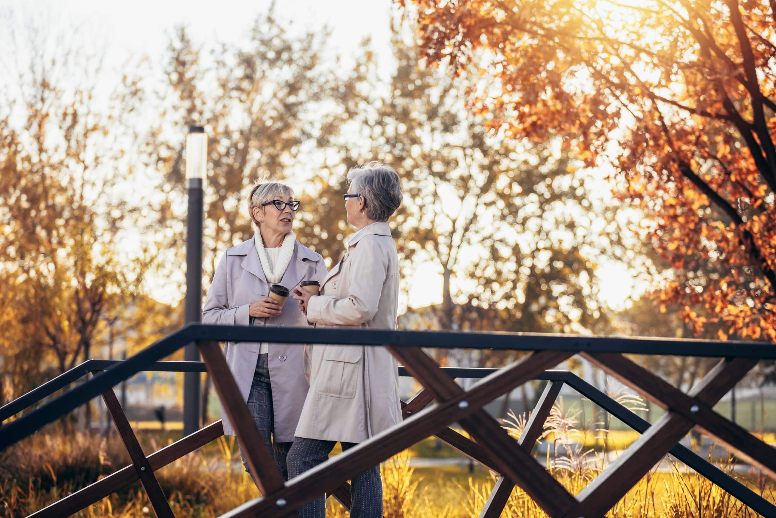 Two 55+ women drinking coffee and walking in a park.