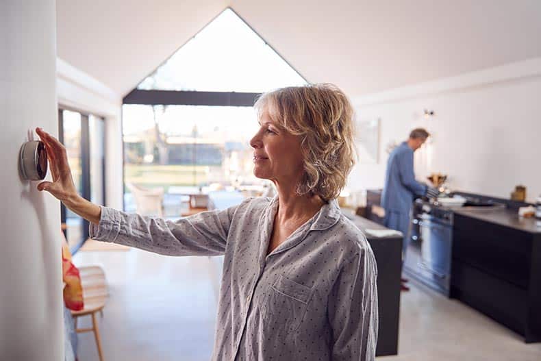 A 55+ woman adjusting a wall mounted digital thermostat at home.